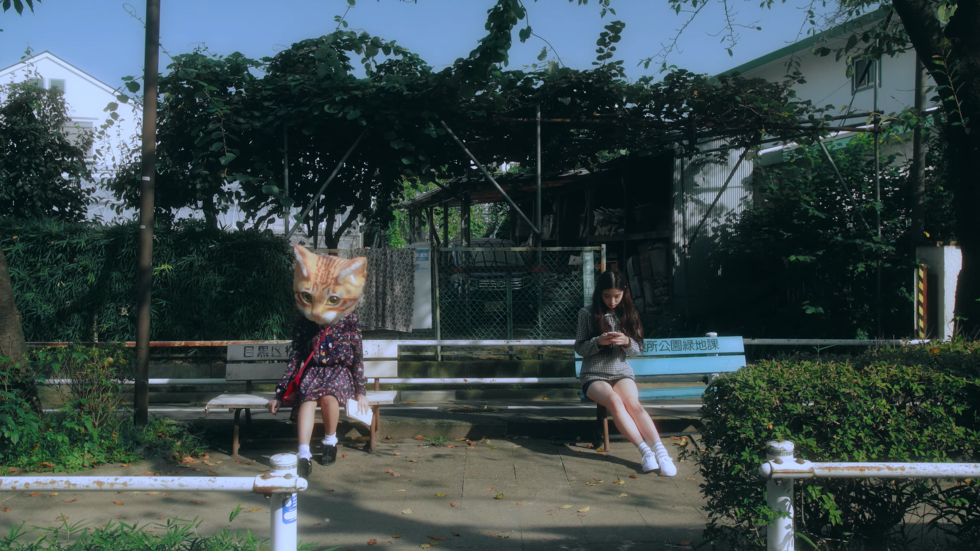 Heejin sitting on a bench, looking at her phone, from the 'Around You' music video. Hyunjin is sitting on a separate bench next to her, wearing a large orange cat head.